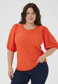 Orange texturerad blus med puffärmar, som har ett diamantmönster. Stylerad med mörka jeans, vilket framhäver en rund halsringning och en böjd nederkant.