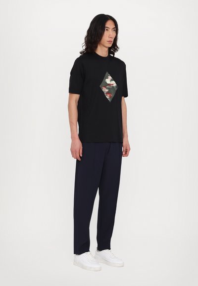 Tricou negru cu mânecă scurtă, cu imprimeu în formă de diamant cu camuflaj; asortat cu pantaloni largi bleumarin și adidași albi.