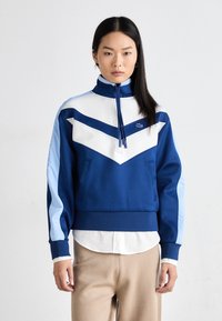 Giacca zip-up blu e bianca con colletto alto, caratterizzata da un design a zig-zag, taglio sportivo e tessuto morbido. Maniche blu solide con accenti più chiari.