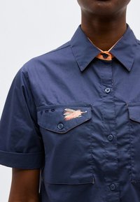 Σκούρο μπλε κοντομάνικο πουκάμισο με γιακά και ροζ-μπεζ κεντημένο φτερό στο καπάκι της αριστερής τσέπης στο στήθος.