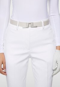 Białe spodnie o gładkiej fakturze i dopasowanej sylwetce, zestawione z jasnoszarym paskiem z metalową klamrą w kształcie litery J oraz białą koszulą.