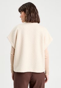 Haut en polaire de couleur crème avec un design boxy, des manches courtes et une texture douce, superposé sur une chemise à manches longues côtelées dans une couleur claire.