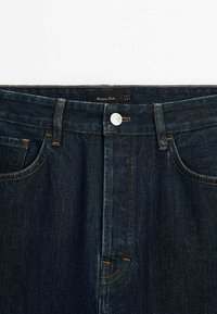 Jean en denim bleu foncé avec bouton visible, passants pour ceinture et poches avant, présentant une couture contrastée et une étiquette noire à l'intérieur de la ceinture.