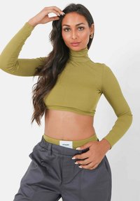 Olijfgroen, geribbeld, langmouw crop top met hoge hals, gecombineerd met donkergrijze broek met elastische tailleband en zichtbaar merkembleem.