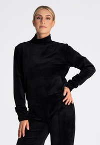 Czarny, długi sweter z długim rękawem z wysokim kołnierzem, wykonany z miękkiego materiału welurowego. Posiada luźny krój i marszczone mankiety.