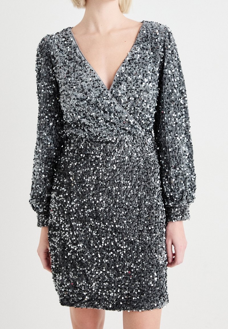 Femme portant une robe portefeuille à manches longues en sequins argentés avec un décolleté en V et une taille froncée.