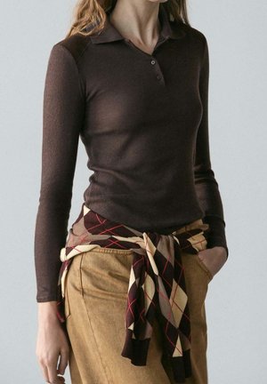 Femme portant un polo marron à manches longues avec un pull à motifs noué autour de la taille et un pantalon beige, debout avec une main dans la poche.