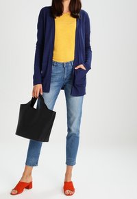 Marinefarbener Cardigan, gelbes Hemd, hellblaue Jeans, rote Blockabsatzsandalen und eine schwarze Tragetasche mit Ausschnitten an den Griffen.