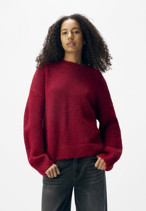 Rode gebreide trui met een losse pasvorm, wijde mouwen en ribbelmanchetten, gecombineerd met donkergrijze broek. Zachte textuur en effen kleur.