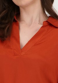 Roest-oranje blouse met een kraag, v-hals en een licht gestructureerde stof. Het ontwerp heeft een gladde afwerking en zachte draping.