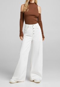 Brauner gerippter Rollkragenpullover mit kalten Schultern, kombiniert mit weißen weit geschnittenen Jeans mit Knopfverschluss und minimalistischem Design.