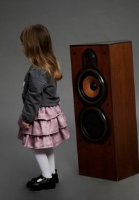 Cassegna in legno con due griglie circolari nere e dorate accanto a un bambino in gonna rosa a strati, felpa grigia e scarpe nere.