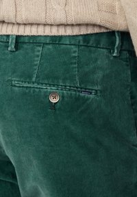 Pantalon en velours côtelé vert avec de fines rayures verticales, doté d'une poche arrière, fermeture à bouton et d'une étiquette contrastée. Détail du tissu texturé visible.