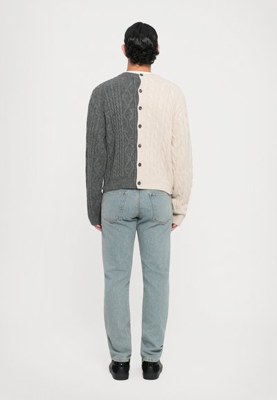 Stickad cardigan med en färgblockerad design i mörkgrå och krämfärgade sektioner, strukturerade kabelmönster och svarta knappar längs ryggen.