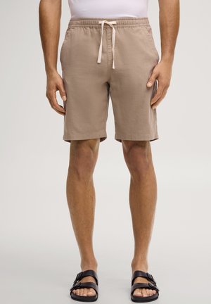 Mężczyzna w beżowych szortach z troczkiem, białej koszuli i czarnych sandałach z klamrą, stoi z rękami luźno ułożonymi po bokach.