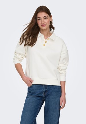 Jeune femme aux longs cheveux bruns, portant une chemise blanche à manches longues avec des boutons jaunes et un jean bleu, debout avec une main dans la poche.