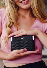 Black textured cardholder with wavy patterns, silver hardware, and a logo on the front, held in hands against a pink top background.