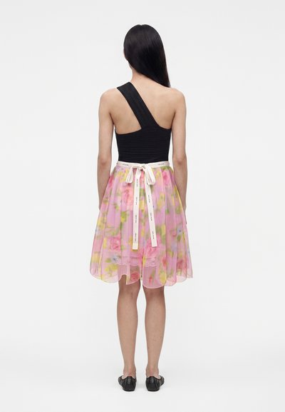 Femme de dos, portant un haut noir asymétrique et une jupe rose à fleurs avec un nœud en ruban blanc, debout devant un fond uni clair.