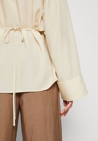 Blouse beige clair avec taille à cordon, manches longues et ourlet arrondi. Le tissu semble lisse avec un léger éclat. S'associe avec un pantalon marron.