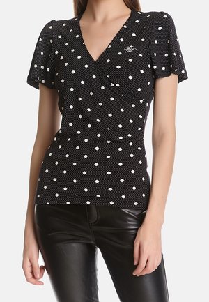 Czarna bluzka kopertowa z krótkim rękawem w białe kropki, noszona przez stojącą osobę, w połączeniu z czarnymi skórzanymi spodniami.