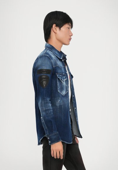Junger Mann im Profil, der eine ausgebleichte blaue Jeansjacke mit schwarzen Patches an den oberen Ärmeln und dunklen Hosen trägt.