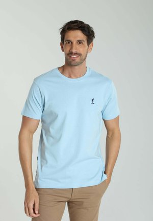 Homme aux cheveux foncés portant un t-shirt bleu clair à manches courtes et un pantalon beige, debout avec une main dans la poche devant un fond uni.