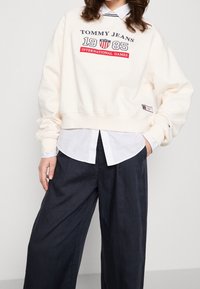Kremowa bluza z logo w granatowym i czerwonym kolorze, krótki krój, ściągacze na mankietach, noszona na białej koszuli z kołnierzykiem, zestawiona z granatowymi szerokimi spodniami.