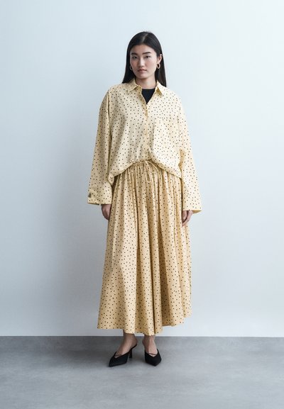 Γυναίκα που στέκεται μπροστά σε απλό τοίχο, φοράει κίτρινη μπλούζα με πουά και μακριά φούστα στο ίδιο μοτίβο, μαύρες γόβες, με τα χέρια χαλαρά δίπλα στο σώμα.