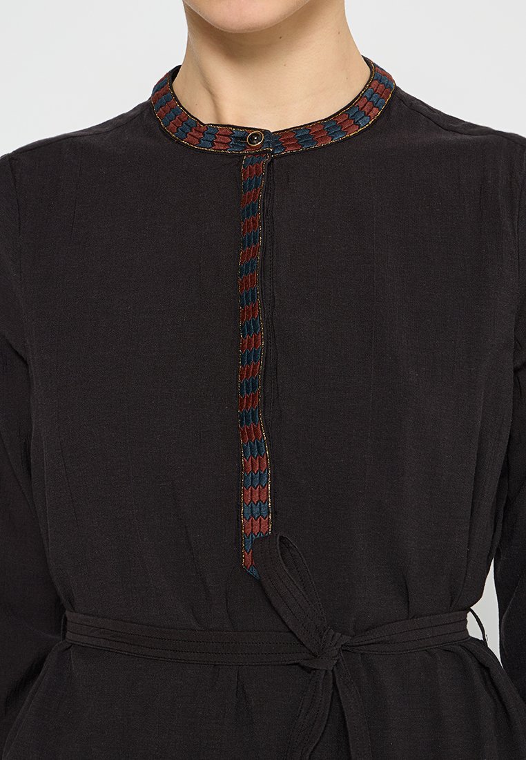 Schwarzes Kleidungsstück mit rundem Ausschnitt, das ein geflochtenes Muster in Rot und Blau aufweist, einen Knopfverschluss hat und mit einem Taillengurt für eine anpassbare Passform ausgestattet ist.