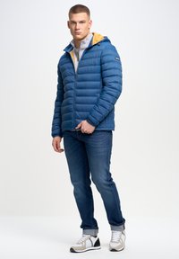 Blå polstret jakke med hætte, med gul foer, lynlås lukning og vandrette sømme. Båret over en grå sweater og blå jeans.