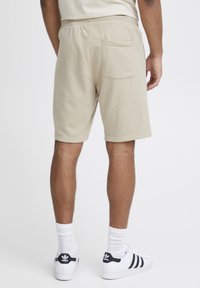 Shorts en coton beige avec une ceinture élastique, des poches latérales et arrière, et une coupe décontractée. Portés avec des chaussettes blanches et des baskets à accents noirs.