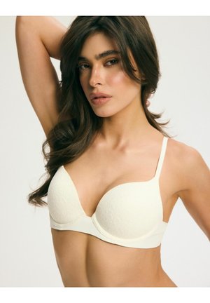 Kobieta o długich ciemnych włosach ubrana w biały teksturowany biustonosz, pozująca z uniesioną jedną ręką na jasnym, jednolitym tle.