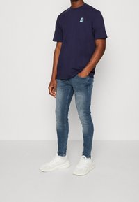Camiseta de manga corta azul marino con un logo azul claro, combinada con jeans ajustados azul claro y zapatillas blancas. Atuendo simple y casual.
