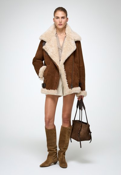 Braune Wildlederjacke mit Schafslederkragen und -manschetten, kombiniert mit cremefarbenen Shorts und knielangen braunen Stiefeln. Das Model hält eine braune Tasche.