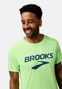 Camiseta deportiva de color verde claro con un gran logo azul de "BROOKS" y un gráfico estilizado, con mangas cortas y un ajuste casual.