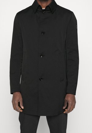 Homme portant un manteau noir boutonné avec un col, des manches longues et des poches latérales, debout devant un fond clair uni.