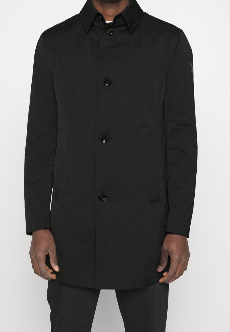 Homme portant un manteau noir boutonné avec un col, des manches longues et des poches latérales, debout devant un fond clair uni.