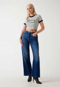 Szürke, rövidített póló fekete szegéllyel és "Guess" logóval, magas derekú, széles szárú kék farmernadrággal és fekete magassarkú csizmával párosítva.