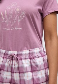 Μωβ βαμβακερό T-shirt με floral σχέδιο και το κείμενο "Ώρα για άνθιση", συνδυασμένο με καρό σορτς σε μοβ και λευκούς τόνους με ελαστική μέση και κορδόνι.