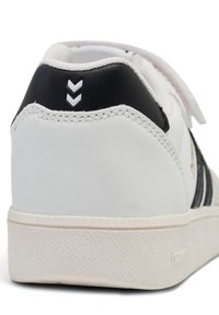Sneaker bianco con accenti neri, caratterizzato da una tomaia in pelle liscia, suola in gomma testurizzata e cinturino in velcro. Dettaglio a chevron notevole sul tallone.