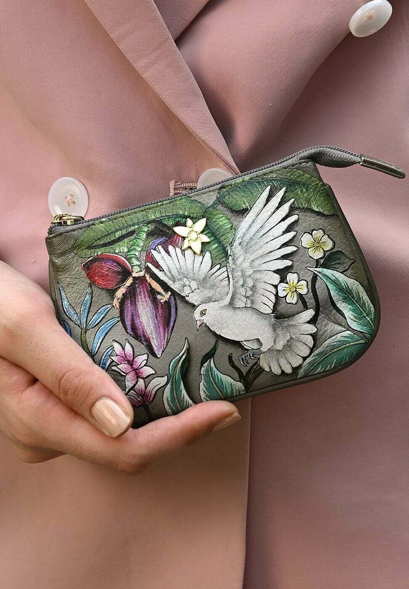 Grijze leren portemonnee met ingewikkelde bloemen- en vogelontwerpen, gouden rits en een gestructureerde afwerking. Gehouden tegen een roze stoffen achtergrond.