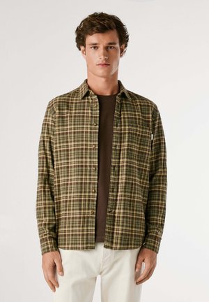 Camicia flanella a quadri verde e marrone con maniche lunghe, chiusura a bottoni e taschino sul petto, indossata sopra una maglietta marrone scuro.