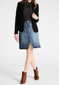 Woman wearing a black blazer, brown polka dot blouse, knee-length blue denim skirt with front slit, and brown suede ankle boots.