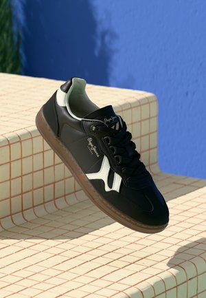 Sneaker noir avec des accents blancs et une surface texturée. Dispose d'un bout rond, d'un design à lacets et d'une semelle en caoutchouc naturel. Logo de la marque visible.