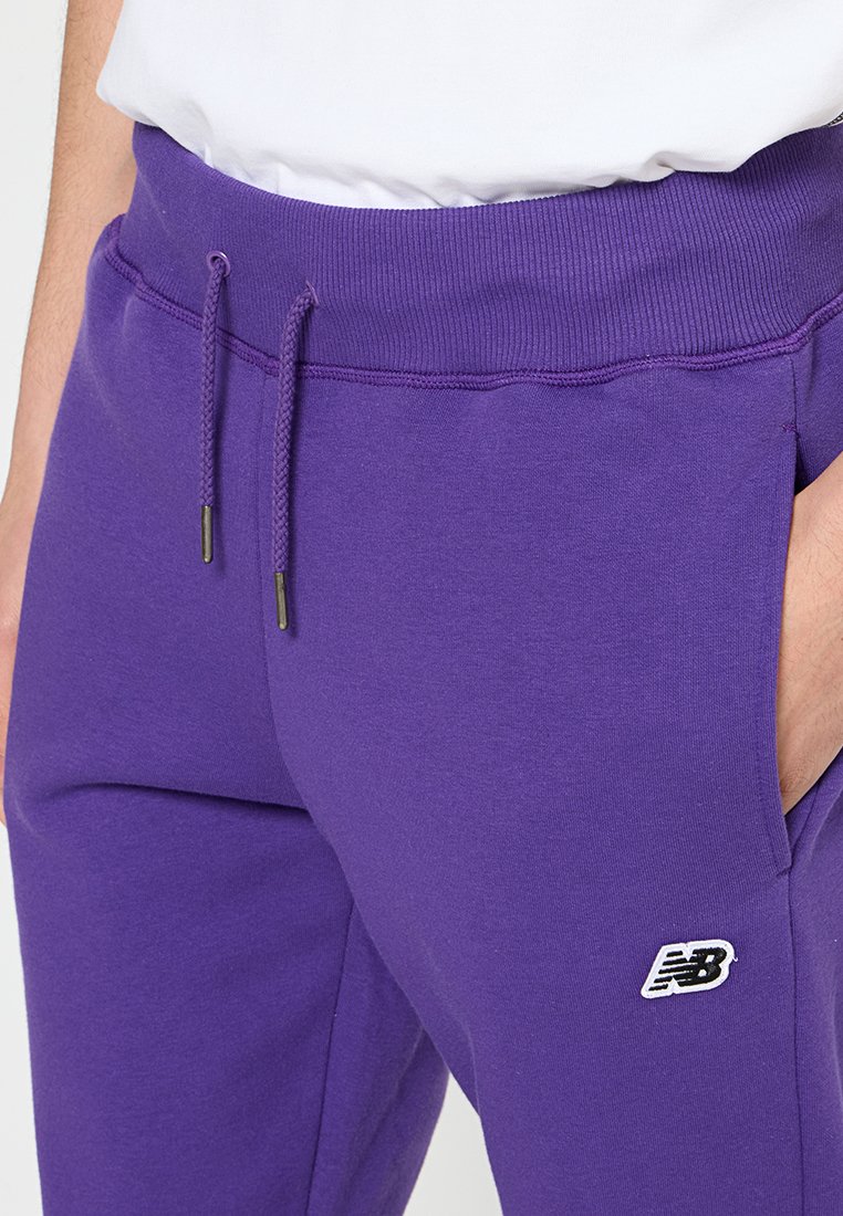 Pantaloni da tuta viola realizzati in tessuto misto cotone, con vita regolabile mediante cordoncino, tasche laterali e un piccolo logo bianco di New Balance sulla coscia.