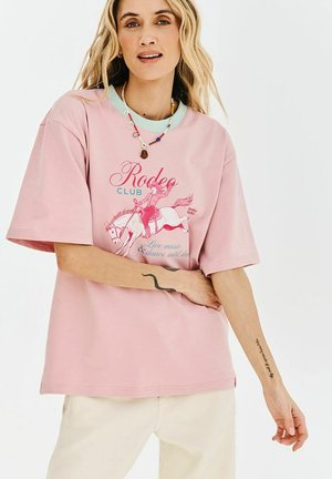 Młoda kobieta o falowanych blond włosach nosi różowy, luźny T-shirt z grafiką jeźdźca rodeo, białe spodnie oraz koralikowy naszyjnik, ukazując tatuaże na ramionach.