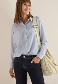 Camicia a righe blu chiaro con bottoni e maniche lunghe, abbinata a jeans scuri con cuciture a contrasto e una borsa a tracolla beige.