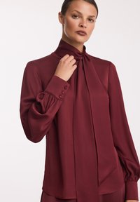 Burgundowa bluzka z wysokim kołnierzem, detalem w postaci kokardki i bufiastymi rękawami. Wykonana z gładkiego materiału z guzikami na mankietach.