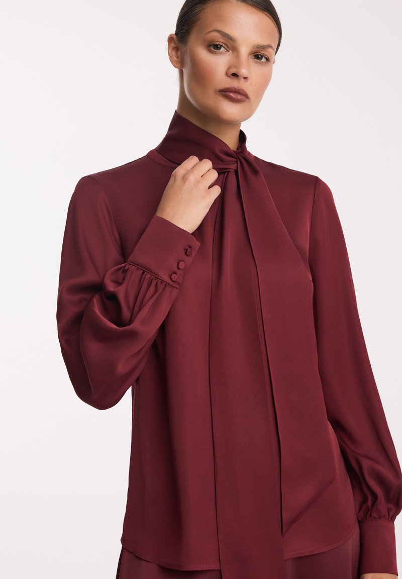 Burgundowa bluzka z wysokim kołnierzem, detalem w postaci kokardki i bufiastymi rękawami. Wykonana z gładkiego materiału z guzikami na mankietach.