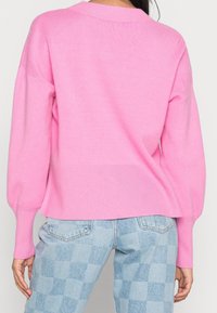 Różowy sweter z dzianiny z prążkowanym dołem i mankietami, o luźnym kroju i szerokich rękawach, zestawiony z jasnoniebieskimi jeansami w kratkę.